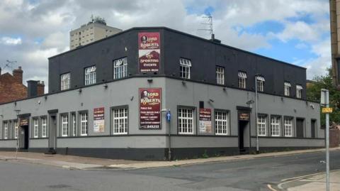 A grey building with a darker grey roof, it has signs on and is on the street corner 