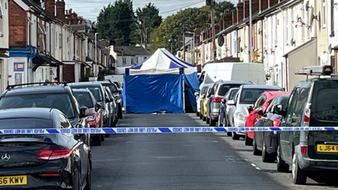 A blue police tent in a residential street, with a police cordon in the foreground