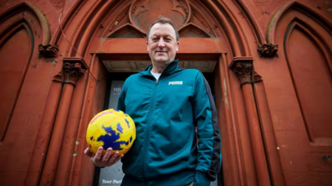Tony stands outside his community hub in Birkenhead with a football in hand. He is smiling and wears a teal green matching tracksuit.