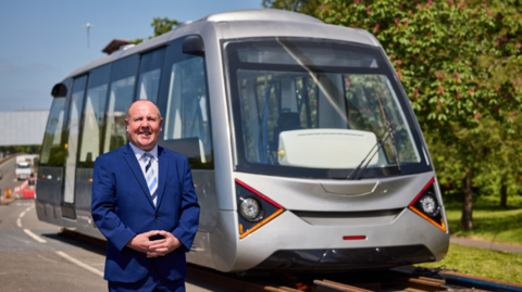 A bald man in a blue suit and stripy blue tie smiles at the camera while standing in the road in front of a very light rail vehicle, which is silver and looks like a tram on rails. It has a large front window with triangular lights on the front