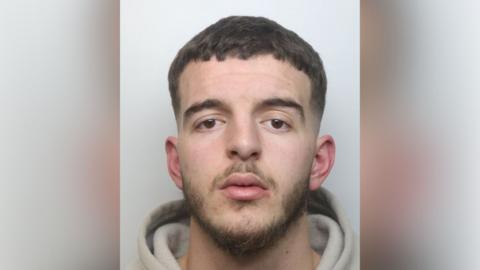 Rebian Bilalaj with short brown hair and beard staring at the camera and wearing a light-coloured hoodie in a police custody photo.