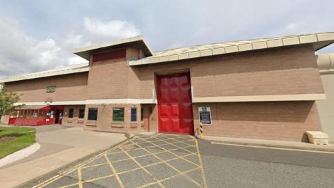 Front of Holme House prison. The low rise building is of brick, with a tall red door and a hatched area in front of it. To the left are some windows and what appears to be a security booth. 