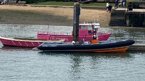 A lifeboat docked in water by a jetty. It is navy blue and orange, and reads "Hamble Lifeboat" on the side. There are two other red boats docked behind it.