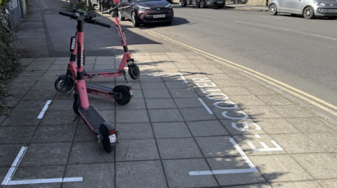 Three red electric scooters parked inside a white parking bay space on the pavement which has the words 'E-SCOOTER' painted by it. 