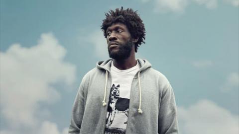 Stormzy as 'Tenzman' in Big Man. Wearing an afro wig and a white t-shirt and grey hoodie.