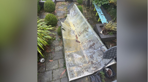 A ripped off rooftop shown damaged and dirty in a yard. There's debris everywhere and pots of plants are scattered.