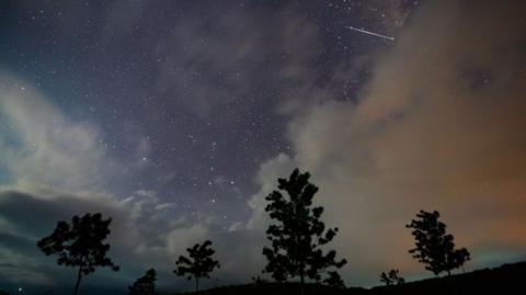 A meteor is seen streaking across the night sky framed by clouds and trees