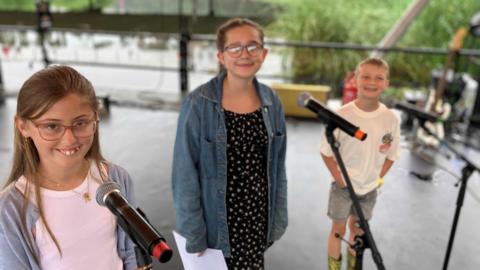 Two 11 year old girls and a 7 year old boy stage on a festival stage with microphones in front of them