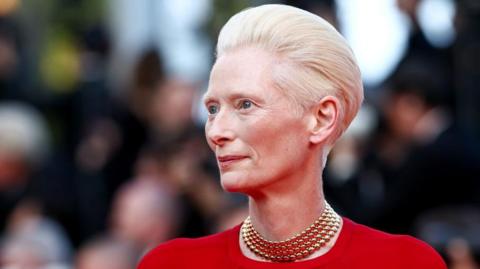 Actress Tilda Swinton, who has short, blonde hair, is dressed in red and wearing a gold necklace. She is smiling and looking to the left.