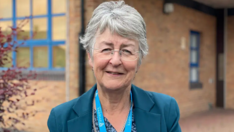 A woman with grey hair and glasses is in a blue jacket in front of a brown building, which is blurred. She is wearing a blue lanyard.