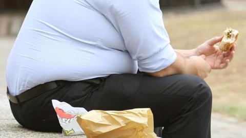 An overweight man sitting on a bench/wall eating a sandwich. 