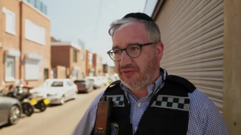 A man white a grey beard and grey hair talks to the camera. He is wearing a black kippah, black glasses, black vest and blue and white chequered shirt. Behind him is a row of houses and a street full of parked cars.