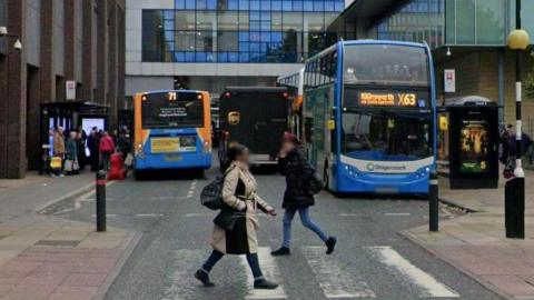 Two women cross a pedestrian crossing on Blackett Street Newcastle. There are two Stagecoach buses in the background and people waiting at a bus station. Part of Eldon Square shopping centre is in the background.