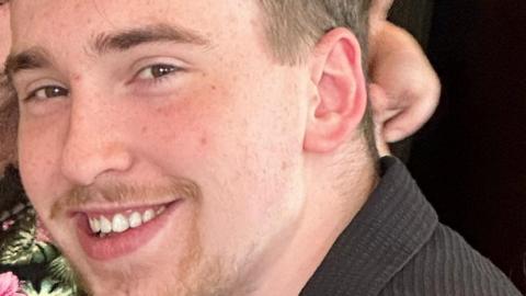 Close-up of a young man with light brown hair and a goatee who is smiling. He is black top with a collar.