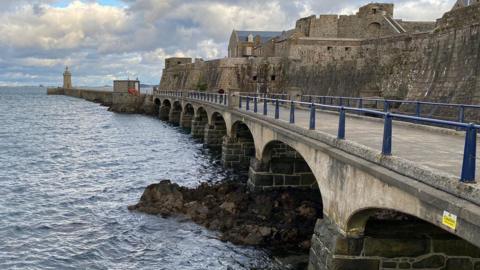 an old stone bridge sits over seawater, alongside a stone castle and leading towards a lighthouse.