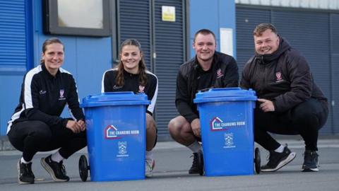 Staff and volunteers at the foundation wearing sports clothing and crouched behind the blue Changing Room bins.