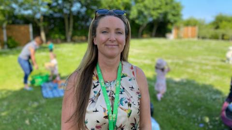Zara Betts, in a cream dress with birds and wearing a green lanyard. Her hair is long ad is wearing sunglasses on her head. There is grass on the ground behind her. The background is blurred. There is a person behind her and three children. 
There is a fence and trees. 