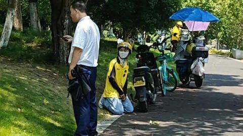 A delivery rider in a yellow uniform kneels next to their bike on the side of a road, as a security guard in a white shirt stands next to them.