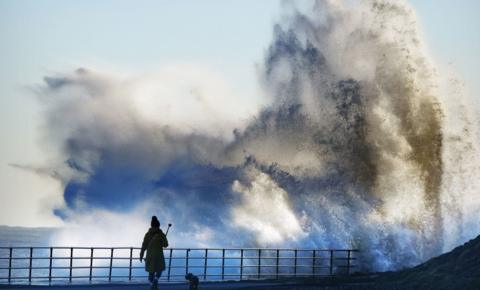 A walker and dog are silhouetted in black against a huge wave striking the sea front at Whitley Bay. The wave is coloured blue at the bottom and white higher up