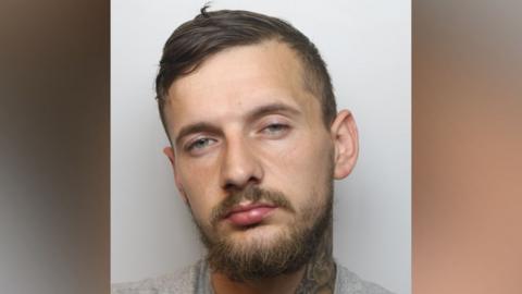 A mugshot of a man set on a blurred background. The man has short hair and a beard, and a neck tattoo is showing.