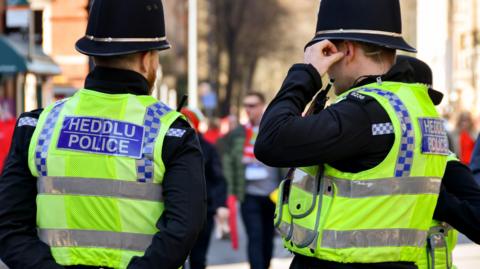 Two police officers stand facing away from the camera. They are wearing fluorescent vests with the words heddlu and police on the back and are wearing black helmets. 