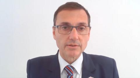 Dr Noble in front of a white background. He is wearing glasses, a dark suit and a tie with red and blue stripes.