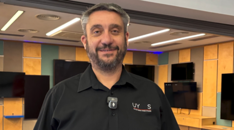A man with grey hair and a beard is wearing a black short-sleeved shirt. He is looking at the camera and more than half a dozen TV screens mounted on a yellow, green and brown wall behind him are switched off.