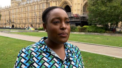 Taiwo Owatemi is wearing a blue, green and white geometric blouse, behind her are the Houses of Parliament