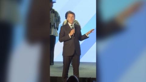 Emmanuel Macron on stage with a mic at Africa Forward summit. He's wearing a dark grey suit and has his hand raised.