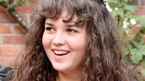 A photo of Maeve. She had brown, curly hair and a fringe covering her forehead, she is looking off to the left and smiling