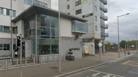 A screengrab of a Google Streetview image of Ballymun Garda Station, a modern white and grey multi-story building with large glass panels at its main entrance. There is a liveried garda van parked between the building and the tall apartment block next door. 