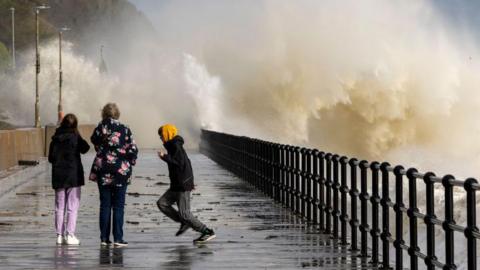 Three people watch as waves hit a promenade, leaving a cloud of white water in the air