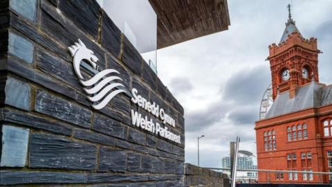 A view of the Welsh Parliament on the left hand side, showing a part of the building made in slate with the parliament's dragon logo emblazoned in white, with the red terracotta Pierhead building on the right.