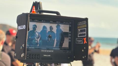 The screen of a film camera which has people in the frame. The camera is black and has different stickers on it, and a small white and orange traffic cone on top of it on the left. In front of the camera are blurred people on a beach. 