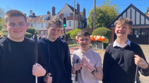 Four boys teenagers who are blind or vision impaired next to a college crossing. Three of them have dark tops and the fourth has a white polo shirt