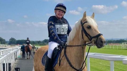 Karen Tidman sits on her horse and smiling. She is wearing dark blue silks, bearing the number 135, and white jodhpurs. She and her horse are on a tarmac path next to a racecourse.