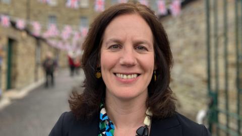 Helen Bishop-Stephens is smiling at the camera. She has shoulder-length dark hair and brown eyes. She is wearing a brightly-coloured top under a black jacket. Behind her there are three people walking down a narrow street under Union Jack bunting.