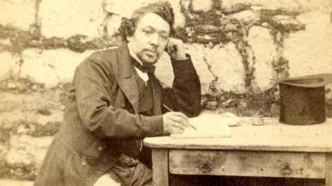 A sepia photograph of James Brown in Victorian gentlemen's clothing sitting at a wooden table with a pen and paper in front of him with the stone walls of Castle Rushen behind him.