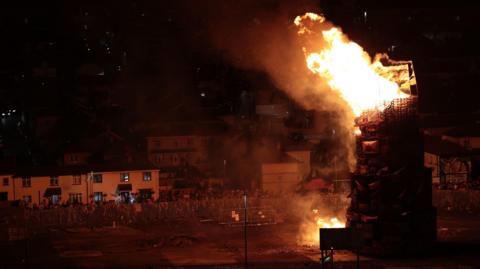 An aerial shot of the bonfire set alight in the bogside of Derry. Flames are ablaze on top of a large tower of pallets with flags and emblems on them. Crowds are standing a few hundred metres away behind a barricade. Houses sit behind the crowds. 
