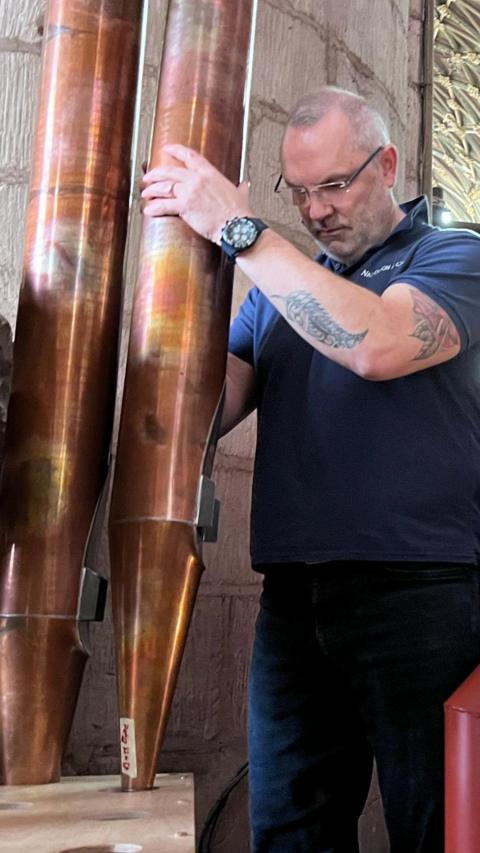 A worker installs or adjusts tall organ pipes inside a cathedral, with scaffolding and wooden organ components around them.