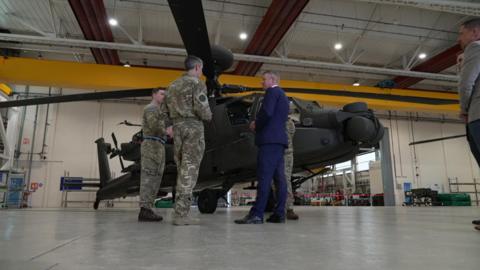 Luke Pollard MP wearing a blue suit stood talking to armed forces personnel in front of an Apache helicopter.