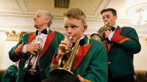 A man with grey hair, a teenage boy with dark hair and a blonde young boy practise their brass instruments before the The National Brass Band Championships of Great Britain. They are wearing green, red and gold uniforms.