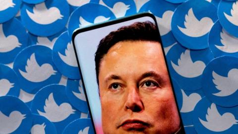 An image of Elon Musk is seen on a smartphone placed on printed Twitter logos