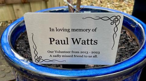 A metal card inside a blue plant pot reads: "In loving memory of Paul Watts. Our volunteer from 2013 - 2023. A sadly missed friend to us all." 