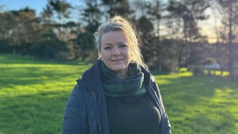 Kirree, a woman with a blonde ponytail, she smiles and wears a scarf and navy puffer jacket as she stands in a field.