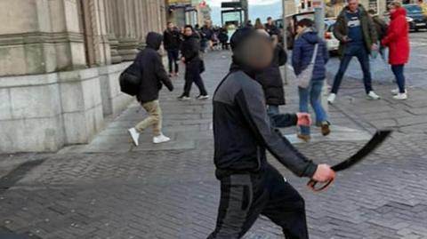 Person holding a large bladed weapon walking across a city centre street, with pedestrians nearby and buildings lining the pavement.