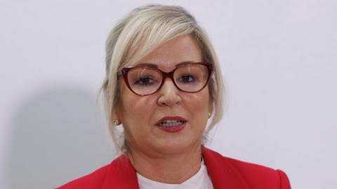 Michelle O'Neill with her hair tied back, she is wearing red cat-eye glasses and a red blazer. 
