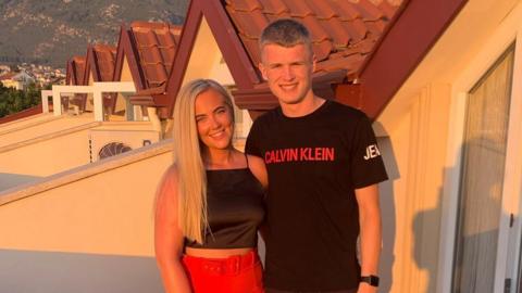 A man and a woman posing side by side for a photograph outside some lodges in a sunny place. The woman is blonde, wearing a black top and red trousers and has a silver bracelet on her right hand. The man is wearing a black top and shorts. 