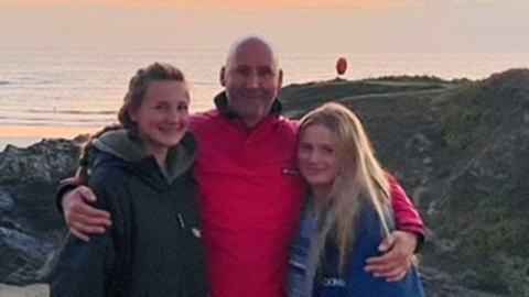 A man and two girls stand on the side of a cliff, which overlooks the beach and the sea.