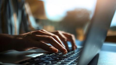A close up of a pair of hands typing on a silver and black laptop keyboard with a window in soft focus behind.